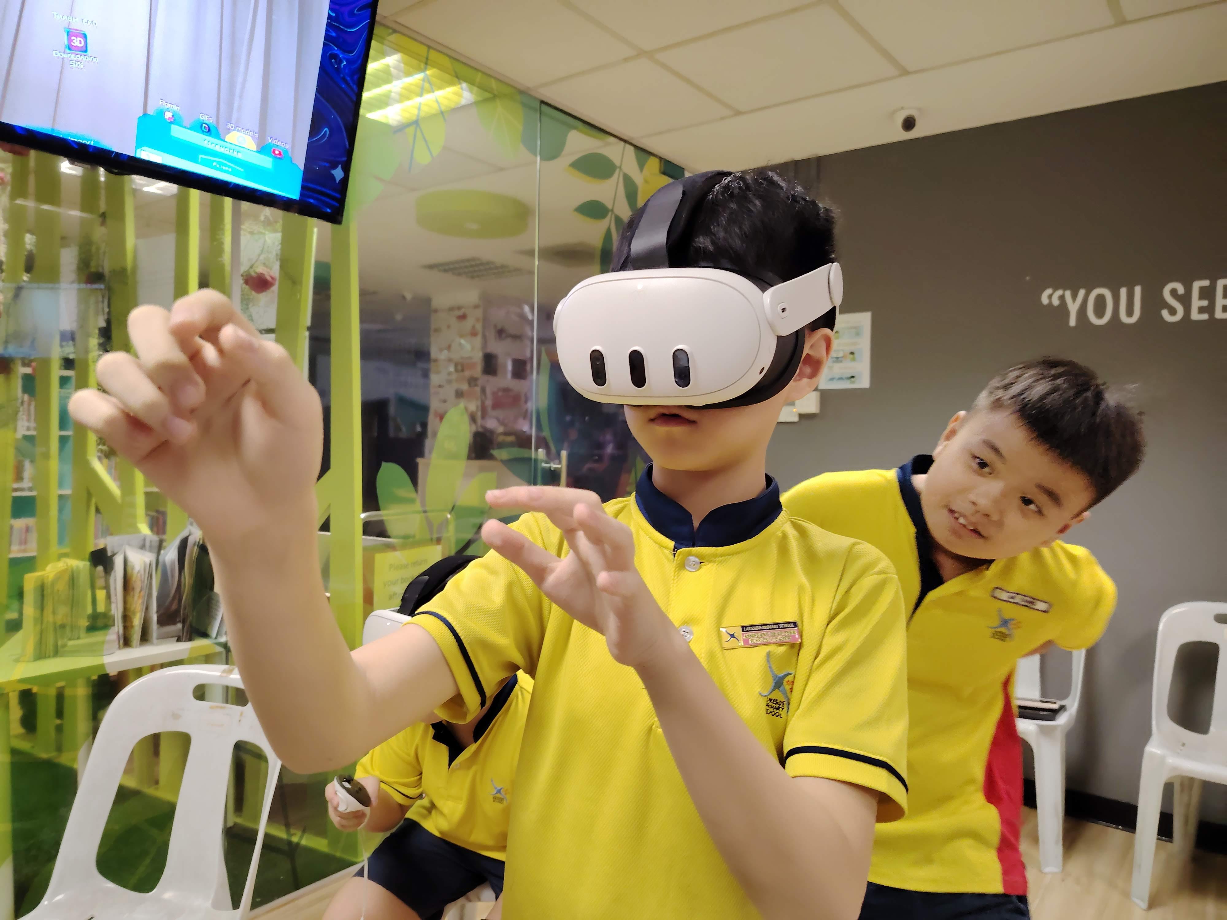 A male student wearing a Meta Quest VR headset raises both hands to interact with the mixed reality environment, while a classmate watches attentively beside him. A screen displaying the VR view is visible in the background.